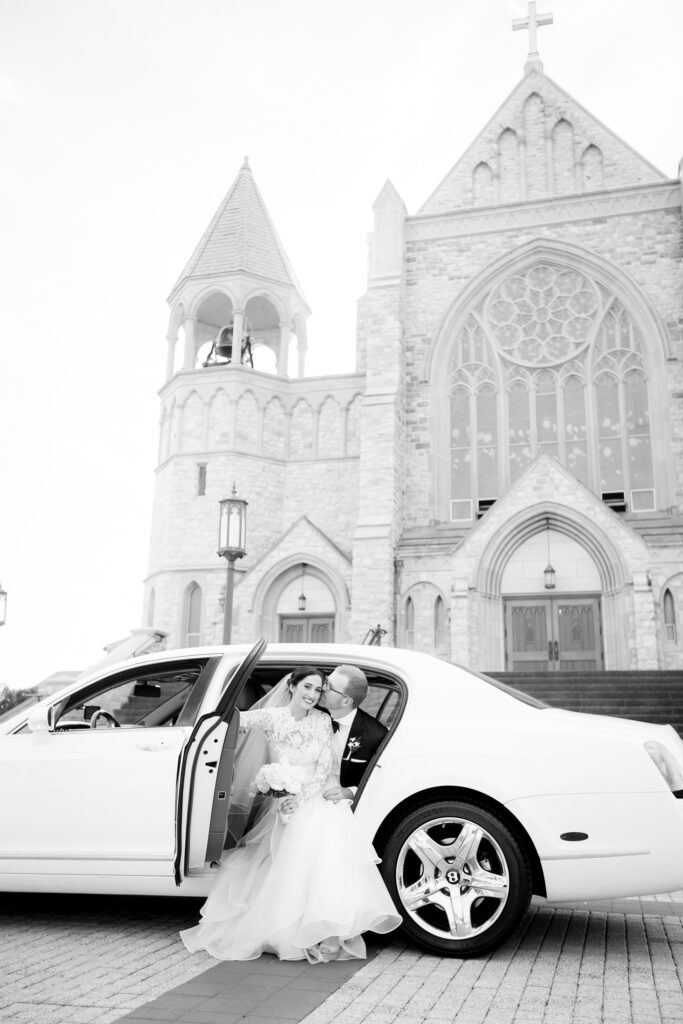 black and white photo of a bride and groom sitting in the back seat of their limousine with their towering church in the background